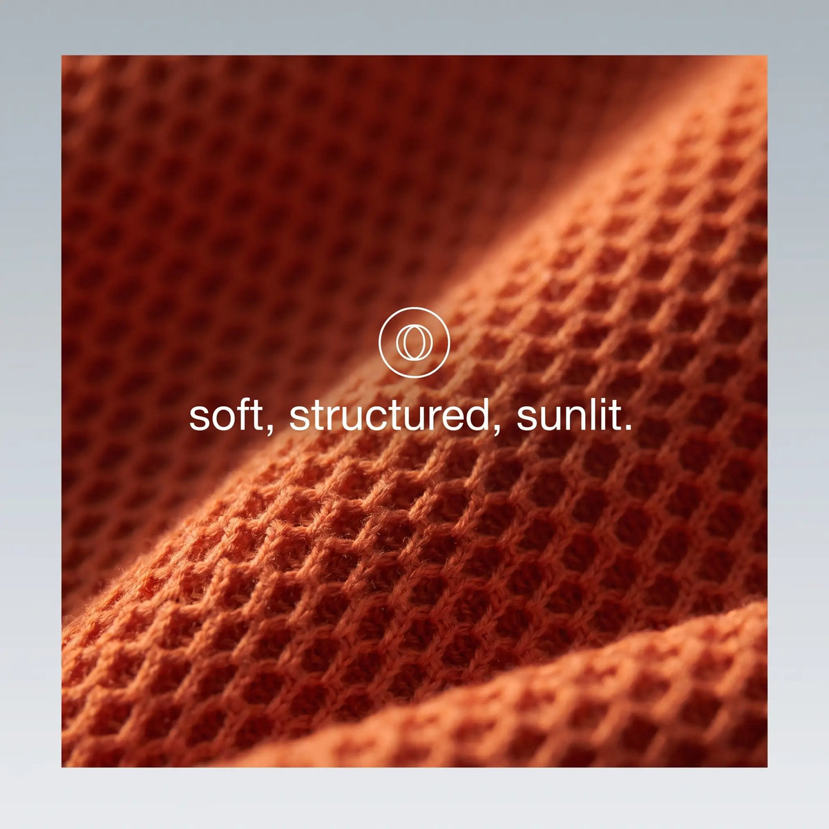 A close-up of textured orange knit fabric with a waffle or honeycomb-like pattern, draped in soft folds under warm light. White text in the center reads 'soft, structured, sunlit.