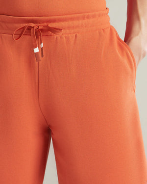 Close-up of a person wearing orange drawstring sweatpants, showing the waistband, tie, side pocket, and part of the matching top against a light neutral background.