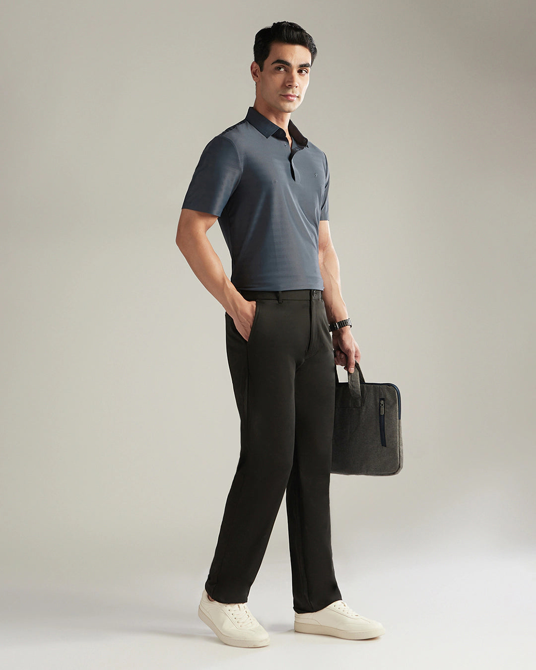 Man wearing a gray polo shirt and black pants, holding a black briefcase against a neutral background