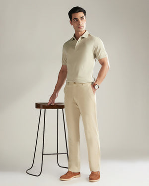 Man wearing a beige polo shirt and pants standing next to a wooden stool on a plain background