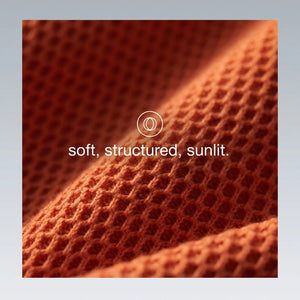 A close-up of textured orange knit fabric with a waffle or honeycomb-like pattern, draped in soft folds under warm light. White text in the center reads 'soft, structured, sunlit.