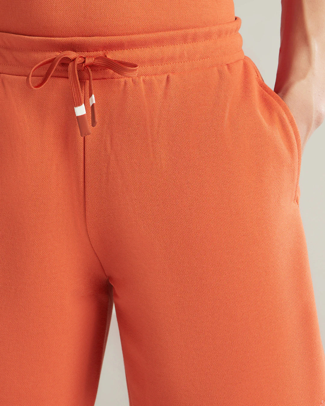 Close-up of a person wearing orange drawstring sweatpants, showing the waistband, tie, side pocket, and part of the matching top against a light neutral background.
