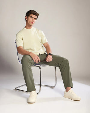 Man sitting on a chair wearing a light-colored sweater, green pants, and white shoes against a plain background