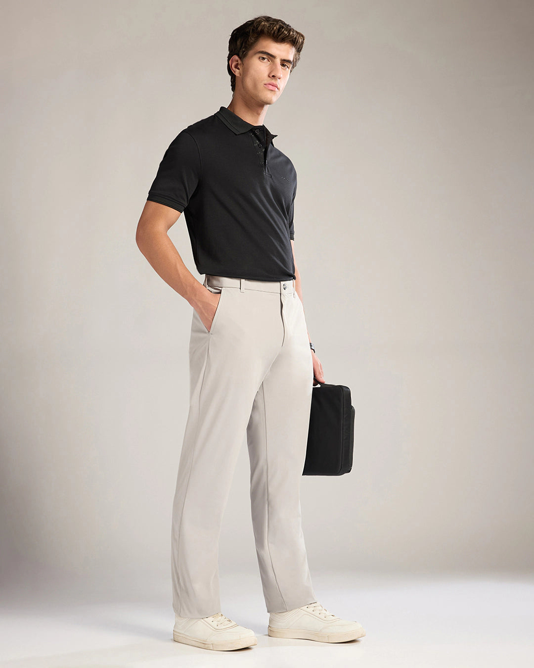 Man wearing a black polo shirt and light gray pants holding a black briefcase against a plain background