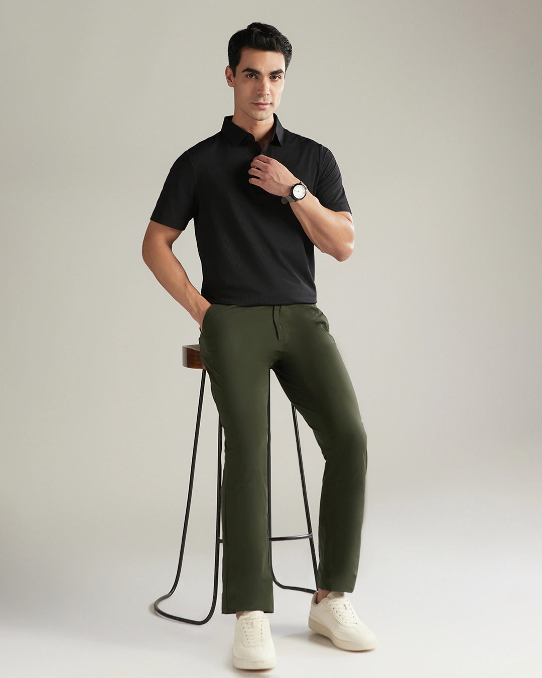 Man wearing a black polo shirt and green pants standing next to a stool on a plain background